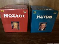 Front view of two box sets, one red labeled Mozart The Masterworks with portrait of Mozart and one blue labeled Haydn The Masterworks with portrait of Haydn, both stating 40 CD box sets.