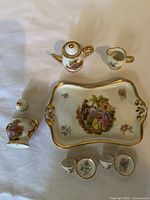 Top view of the miniature porcelain tea set showing the tray with scenic floral image, teapot, creamer, sugar bowl, and two cups with saucers all with gold trim.