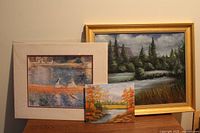 Photo showing two art pieces, one large framed painting of a lake/mountain/evergreen scene in a gold-tone frame right behind a fall landscape painting on canvas.