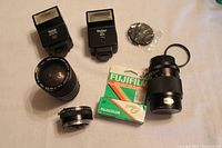 Two Vivitar flashes, lenses, teleconverter, 110 film roll, and lens cover arranged on a surface