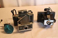 Three vintage cameras displayed on beige fabric surface, showing front and side profiles including the damaged flash on Brownie Hawkeye