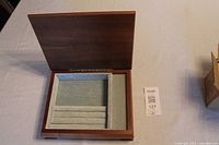 Open wood jewelry box with light green velvet-like lining showing compartments and ring rolls inside