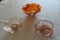 Photo showing three vintage glass pieces: a peach iridescent ruffled bowl, a pink divided candy dish with three compartments, and a marigold-colored Carnival Glass bowl with a ruffled edge and star pattern base.