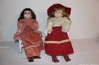 Two soft body porcelain face dolls seated in rattan chairs wearing vintage-style velvet dresses with lace