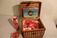 Full view of the wicker picnic basket opened showing the disposable cups, plates, napkins and an insulated bag in original wrapping inside the basket.