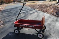 Side and angled view of the red Radio Flyer wagon showing the wagon bed, wheels, and pull handle on pavement outdoors with some fallen leaves nearby.
