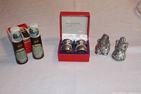 Photo of three salt and pepper shaker sets including Kirk Stieff Santa & Mrs. Claus, Garatino Tre Spade pewter/wood set, and retro 'Mother' and 'Dad' wooden ceramic set. Shakers displayed on a table showing various sizes and styles.