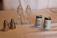 Three pairs of salt and pepper shakers shown together on a table: a clear crystal cut Bohemian pair, a floral ceramic pair with metal tops, and a tarnished silver pair.
