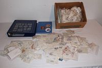 Photo showing a cardboard box filled with small packets of stamps, a dark blue binder with stamp pages, a blue stamp book, and assorted loose paper packets with stamps spread out.