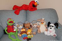 Group shot of all nine stuffed animals on a sofa including Elmo, Kermit Frogtographer, Bert & Ernie, Casey Jones bears, bunny, and dog with sunglasses