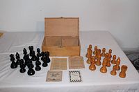 Full setup of chess pieces arranged on table with open wooden box behind and instruction sheets in front