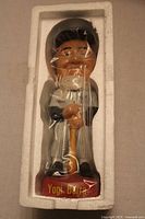 Front view of Yogi Berra bobblehead in packaging showing figure wearing New York Yankees uniform, holding bat, standing on base labeled 'Yogi Berra'.