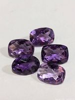 Photo shows five faceted amethyst gemstones in various oval and cushion shapes, with rich purple hues and checkerboard faceting, arranged closely on a white background.