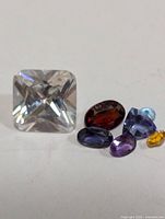 Photo showing six assorted faceted gemstones including a large clear square stone and five smaller colored stones in red, purple, blue and yellow.