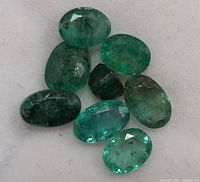 Eight oval cut green emerald gemstones displayed on a white surface, showing color and inclusion details.