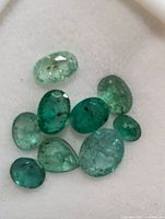 Close up of nine genuine emerald gemstones of various sizes and green hues on white padded background.
