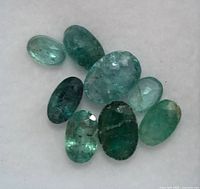 Top-down photo of nine faceted oval emeralds varying in green shades, showing internal inclusions.
