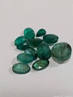 Close-up photo of eleven genuine emerald gemstones in various shapes and sizes, placed on a white surface.