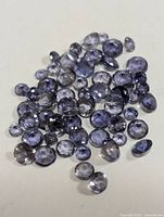 Top view of approximately 48 loose round faceted iolite gemstones with blue-violet tones spread on a white surface.