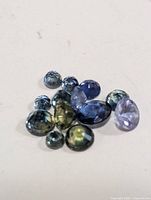 Loose genuine sapphire gemstones in mixed colors, blue, green, purple, faceted, oval and round shapes, grouped on white background.