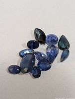 Photo shows eleven loose faceted sapphire gemstones in various cuts and hues of blue on a plain background.