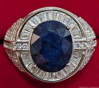 Front view of the men's silver ring featuring an oval blue sapphire center stone surrounded by clear baguette stones.
