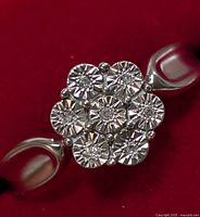Close-up photo of front view of silver ring with 7 round diamonds in cluster floral pattern, on red background.