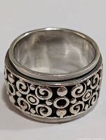Front and side views of a wide silver band ring with intricate raised swirl and dot pattern.