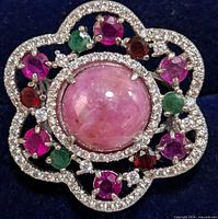 Close-up view of the silver ring showing the detailed arrangement of rubies, emeralds, and small clear stones around a large pink cabochon center stone on a scalloped silver frame.