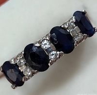 Close-up image of the silver ring showing four faceted dark blue sapphire stones with clear round accent stones in between.