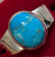 Close-up photo of polished oval turquoise stone bezel set on silver ring band shown against red background, presenting the surface details and veining of the stone and light wear on silver band.