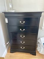 Front view of the black dresser showing five drawers with silver handles and overall shape and finish.