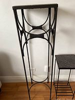Tall black metal plant stand or pedestal table with intricate curved circular design, standing on wooden floor against white wall.
