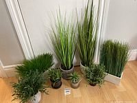 All seven artificial plants arranged in front of a wall on wood floor, showing variety of pots and plant types.