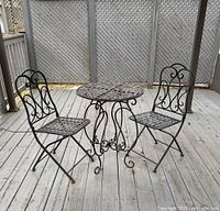 Photo showing the full three piece wrought iron bistro set outdoors on a wooden deck. Includes two matching chairs with scroll backs and a round table with decorative ironwork.
