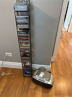 Black metal CD rack filled with several jewel case CDs, placed next to the player on wooden floor against wall.