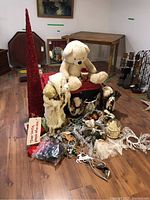 Photo showing large red glittered Christmas tree cone, cream teddy bear holding a mug, Santa figurine, various ornaments, and gift bags spread on floor