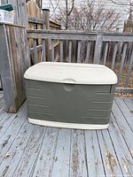 Front view of Rubbermaid deck box showing khaki base and beige lid with latch mechanism, placed on wooden deck with fence in background.