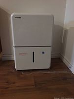Front view of white Toshiba dehumidifier placed on wooden floor against wall, showing water level indicator and Toshiba logo.
