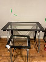 Desk and matching smaller printer table with glass tops, chrome legs, and black frames on a wooden floor against a plain wall.