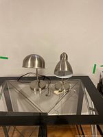 Two metal desk lamps with brushed metal finish on a glass table, one with straight stand and dome shade, the other with a shorter design and a slightly different dome shade.