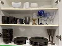 Brown ceramic dishes including bowls, plates of two sizes, mugs, along with white ceramic bowl and pitcher on upper shelf and variety of decorative drinking glasses in different colors and styles on middle shelf.