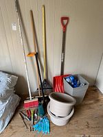 Photo of garden tools: red plastic snow shovel, rakes, push broom, small hand tools on the floor, and beige plastic round planters stacked.