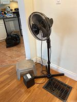 Standing fan, space heater, and grey plastic step stool shown together in room.