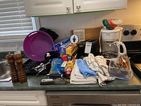 Lot of kitchen items on counter including cutlery in plastic container, wooden salt and pepper mills, plastic purple mixing bowl, packing wraps, and kitchen towels.