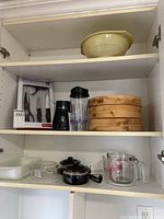Kitchen cabinet with boxed kitchen gadgets, single serving blender, bamboo steamer basket, ceramic strainer, measuring cup, and cookware.