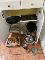 Kitchen cabinet opened, showing various kitchen items including baking trays, roasting pan, metal cooling rack, mixing bowls, glass loaf pan, circular black pan, metal ladle with stand, and cooking booklets on floor.