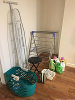 View of entire laundry supply lot showing metal ironing board, indoor drying rack, black foldable stool, teal laundry basket, and laundry detergents.