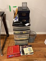 Full view of plastic organizer with drawers, two stacked decorative boxes on top, metal mesh pen holder with office supplies on the side, stapler on top of the organizer, three books on the floor, and a small metal trash bin nearby.