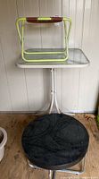 Tall outdoor patio table with rectangular glass top and white metal frame, small folding chair resting on it, and round black table on floor